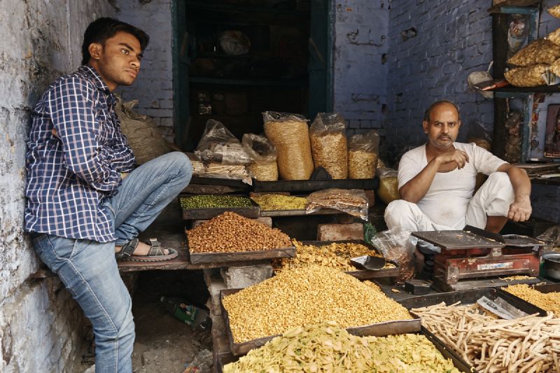 A cereal market in India. In the foreground, a young man leans against the wall of an open shop while another man sits on the ground inside